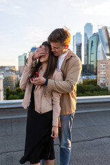 Wall Mural - Handsome man is doing surprise marriage proposal to his beautiful girlfriend on a rooftop. Emotional. Happy loving couple on a romantic date on Saint Valentine's Day. Urban cityscape on background.