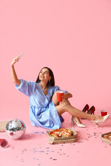 Poster - Young woman with cup, tasty pizza and disco ball taking selfie on pink background