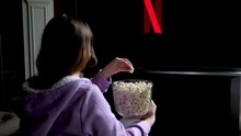 teenage girl sits on floor and watches netflix on big screen of TV screensaver slow motion flashes on black background in red blue in hands of young woman popcorn girl takes it and eats it spellbound