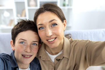 Son and mother at home sitting on the sofa in the living room, looking at the camera, smiling, taking a selfie photo, and talking via video call online.