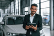 © standret - Holding graphic tablet. Handsome indian man in suit is in the car showroom with the vehicle indoors