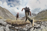 Fototapeta  - Two young hikers walks with light backpacks in mountains. Tourist jumps across the obstacle