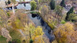 Fototapeta Las - Beautiful view people walking on ground paths near lakes in park with trees with yellow fallen leaves on autumn day. Flying over walking people, ponds, trees with yellow foliage. Aerial drone view.
