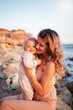 Young mother with her daughter on vacation in the summer on the beach. Mom kissing the baby. Rocky beach, mindful motherhood, baby care, mother's day, first year of a baby's life, parent. Family look