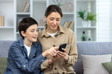 Mother and son sitting together on sofa at home in living room, family looking at phone screen watching video online and choosing shopping in online store hugging and smiling close up.