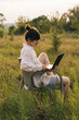 © Ananass - a young woman wearing a shirt works on a laptop while sitting on a chair in the middle of nature