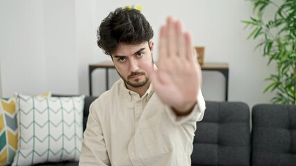 Sticker - Young hispanic man sitting on sofa with serious expression doing stop gesture at home