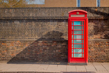 Red Telephone Booth Over A Brick Wall In Cambridge. Old Vintage English Telephone Booth On A Cool Background Casting Shadow.

