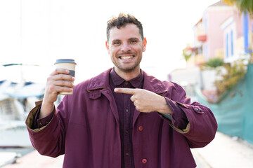 Wall Mural - Young caucasian man holding a take away coffee at outdoors and pointing it