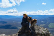 © Ekaterina - Pet owner spends time in nature with German and Australian Shepherds. Hiking in mountains with dogs. Young Caucasian woman sitting on edge of cliff on warm sunny spring day with two dogs.