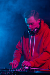 Wall Mural - Cool young hip hop DJ in red hoodie mixing vinyl records on a party in night club. Professional disc jockey plays set in smoke on stage