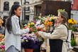 © Asier - Young Latina woman purchasing vibrant flowers and plants from a street vendor's stall, handed by the gardener.
