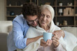 Loving young adult son giving positive sick senior mother help, assist, care, wrapping mom shoulders into plaid, scarf, giving cup of hot drink, hugging, smiling, laughing