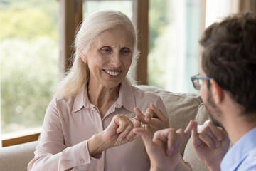 Happy positive old lady with hearing disability speaking sign language to young man, adult son, caregiver, showing hand signs, smiling, laughing, enjoying talk, communication with therapist