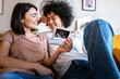 © Daniel - Bliss. Happy and joyful multiracial lesbian pregnant couple looking baby ultrasounds at home relaxing on the sofa.