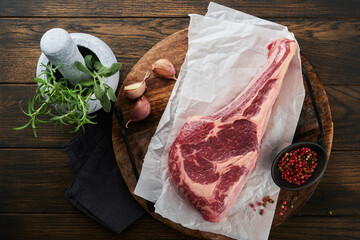 Wall Mural - Fresh tomahawk raw steak. Dry aged raw tomahawk beef steak with herbs and salt on old wooden background. Preparing to grill.  Top view and copy space.