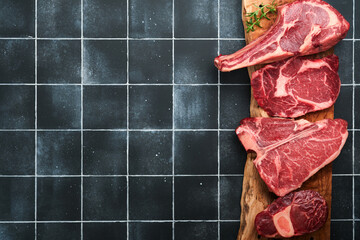 Wall Mural - Raw prime steaks. Variety of fresh black angus prime meat steaks T-bone, New York, Ribeye, Striploin, Tomahawk on black old tiles stone background. Set of various classic steaks. Top view copy space.