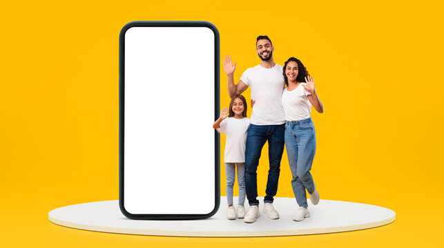 Wall Mural - Glad young middle-eastern family with small girl waving hands near huge phone with blank screen