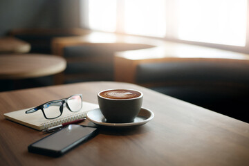 Close-up view, white cup of coffee with smartphone, notebook, pen and eye glasses on wooden table in cafe. Vintage light, blurred background