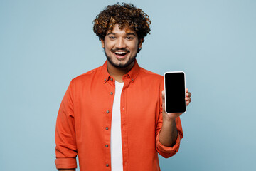 Wall Mural - Young smiling Indian man wears orange red shirt white t-shirt hold in hand use mobile cell phone with blank screen workspace area isolated on plain pastel light blue cyan background studio portrait.