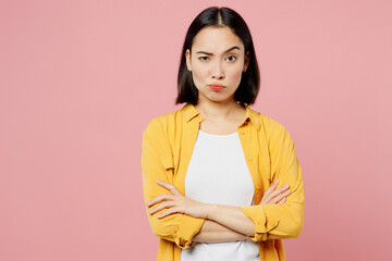 Young displeased sad woman of Asian ethnicity wear yellow shirt white t-shirt hold hands crossed folded look camera isolated on plain pastel light pink background studio portrait. Lifestyle concept.