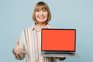 Elderly IT woman 50s years old wear light striped shirt hold use work on laptop pc computer with blank screen workspace area show thumb up isolated on plain pastel blue cyan color background studio