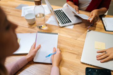 Close-up high-angle view of focused businesswoman holding notebook and pen in hand and write during multicultural professional businesspeople working together, on research plan in boardroom.