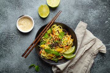 Asian vegetarian noodles with vegetables and lime in black rustic ceramic bowl, wooden chopsticks, cutting board with chopped green onion top view on stone background. Cooking noodles concept