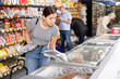 Attentive young woman purchaser choosing frozen fish out of large stock in supermarket