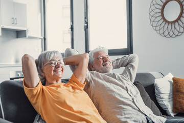 Peaceful middle aged man and woman with closed eyes relaxing on comfortable couch at home, mature family daydreaming together, grey haired wife and husband resting with hands behind head, breathing.