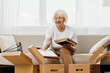 © SHOTPRIME STUDIO - elderly woman sits on a sofa at home with boxes. collecting things with memories albums with photos and photo frames moving to a new place cleaning things and a happy smile. Lifestyle retirement.