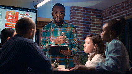 Poster - African american business coach giving presentation in company meeting, holding clipboard, reading report. Employee showing analytics research charts, statistics at night time. Handheld shot.