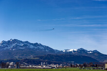 City Of Emmen With Mount Pilatus In The Background And Fighter FA 18 Hornet J-5001 Of Swiss Air Force Up In The Air On A Sunny Spring Day. Photo Taken March 22nd, 2023, Emmen, Switzerland.