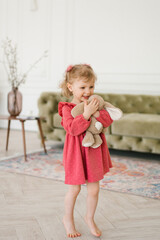 Three-year-old girl in a barefoot dress holds a plush bunny in her hands and hugs him at home in the living room