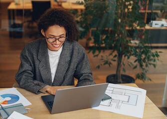 Wall Mural - Smiling woman manager working on laptop in modern coworking while sitting near colleagues