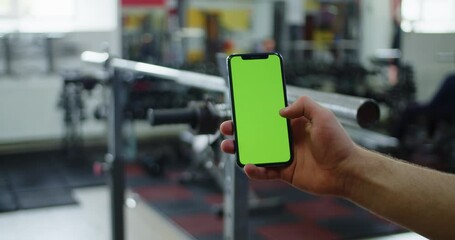 Male hand holding smartphone in gym, fitness app for bodybuilding, green screen.