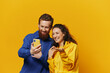 © SHOTPRIME STUDIO - Man and woman couple smiling merrily with phone in hand social media viewing photos and videos, on yellow background, symbols signs and hand gestures, family freelancers.
