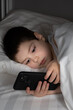 © Zarina Lukash - A 5 year old boy plays games on his phone while lying in bed. Gadgets in bed before bedtime in a child