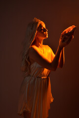 Poster - portrait of beautiful blonde woman wearing a fantasy goddess toga costume with  crystal crown.
Backlit a glowing isolated studio background with  orange silhouette lighting.