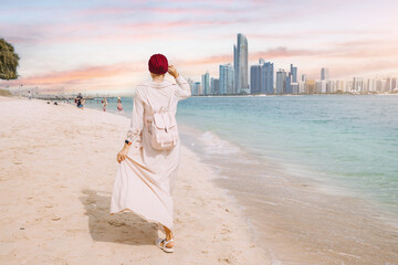 Wall Mural - Despite the size and grandeur of the skyscrapers, the woman seems undaunted as she walks confidently along the shoreline, perhaps lost in thought or simply enjoying the peaceful moment.
