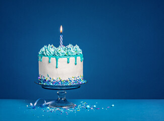 Wall Mural - White birthday drip cake with teal ganache and lit candle over dark blue background