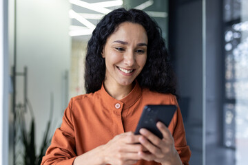 Wall Mural - Beautiful and successful Hispanic woman at work smiling and looking at the phone screen, businesswoman in the office holds smartphone in her hands closeup browses the Internet uses application call