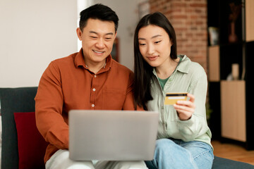 Wall Mural - Smiling korean middle aged man and young woman using credit card and laptop, sitting on sofa in living room interior