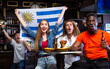 Multiracial Uruguay sports fans, men and women, supporting their favourite team in bar, raising state flag and screaming chants together.