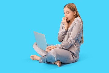 Young woman in pajamas with laptop biting nails on blue background
