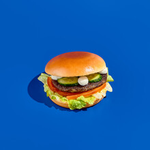 Tantalizing Burger With Feta Cheese, Fresh Cucumbers, Tomatoes, White Sauce, And A Meat Patty On A Blue Background. Contemporary Minimalist Food Photography, Great For Takeout. Square, Low-angle.