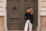 Fototapeta Na drzwi - Calm stylish woman having good fashion clothes walking on street and holding tea or coffee in cup takeaway with good mood. Girl drink coffee, standing near wood green door and waiting for friends.