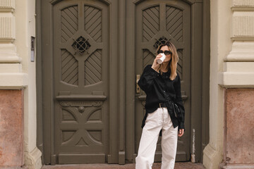Wall Mural - Calm stylish woman having good fashion clothes walking on street and holding tea or coffee in cup takeaway with good mood. Girl drink coffee, standing near wood green door and waiting for friends.