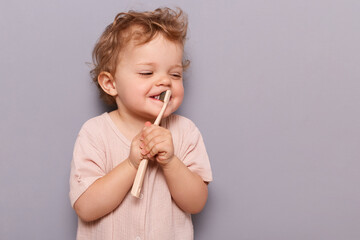 Horizontal shot of happy kid girl brushing teeth, holding toothbrush in hands, trying to do it herself, standing isolated over gray background, copy space for advertisement.