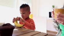 Little Hispanic Boy Having Breakfast With His Mom, He Is Peeling A Banana